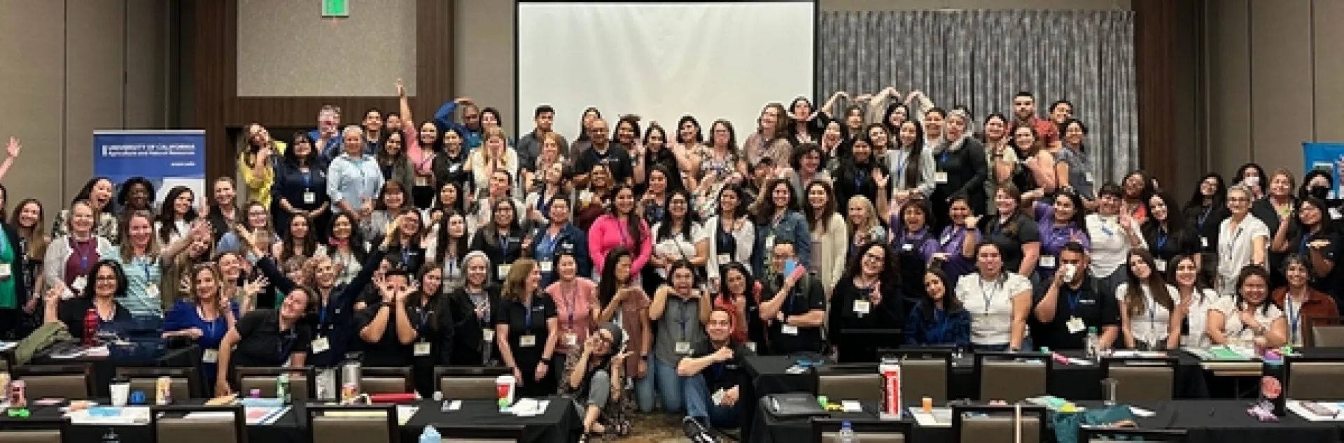 About 100 members of the Community Nutrition and Health network ham it up for a group photo in a conference room.