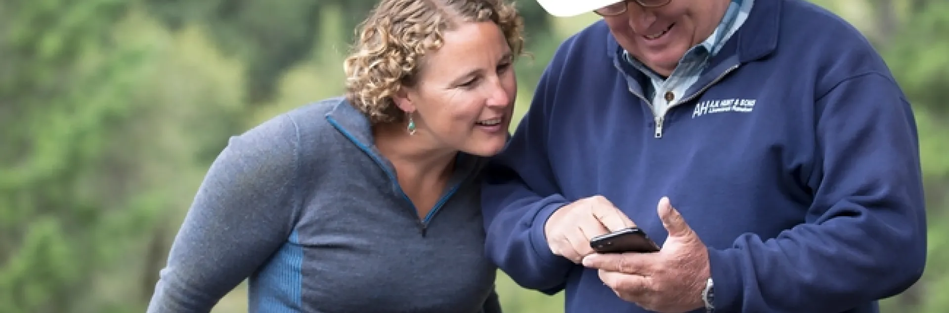 Lenya smiles and looks at whatever a man in a cowboy hat is pointing at on his phone.