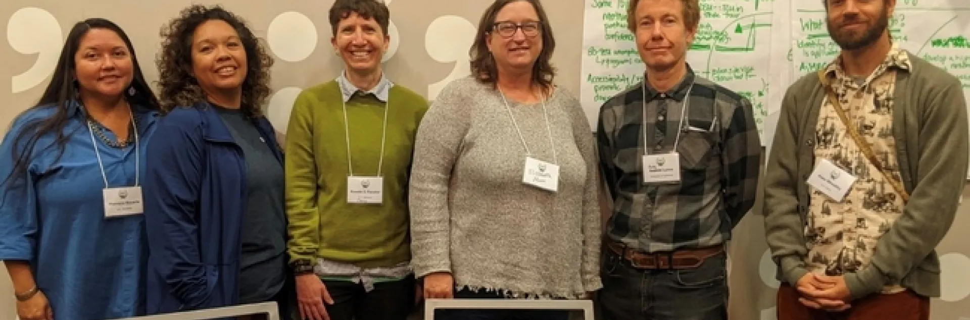 Six people wearing name tags stand in front of a wall that is partly covered in white poster paper with discussion notes written in green ink.