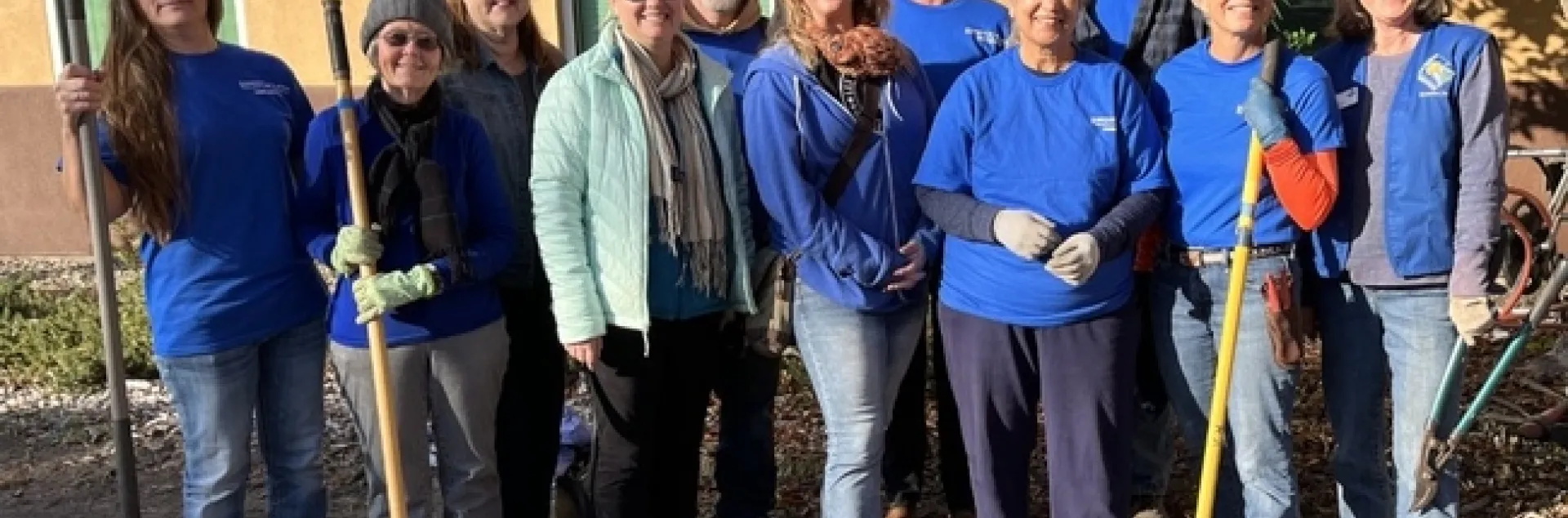 Smiling group of people, standing in two rows while holding gardening tools.