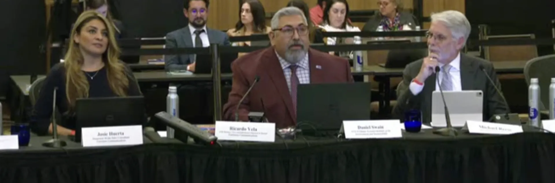 Three people sit at a long table facing the camera. A gallery of spectators sit behind them.
