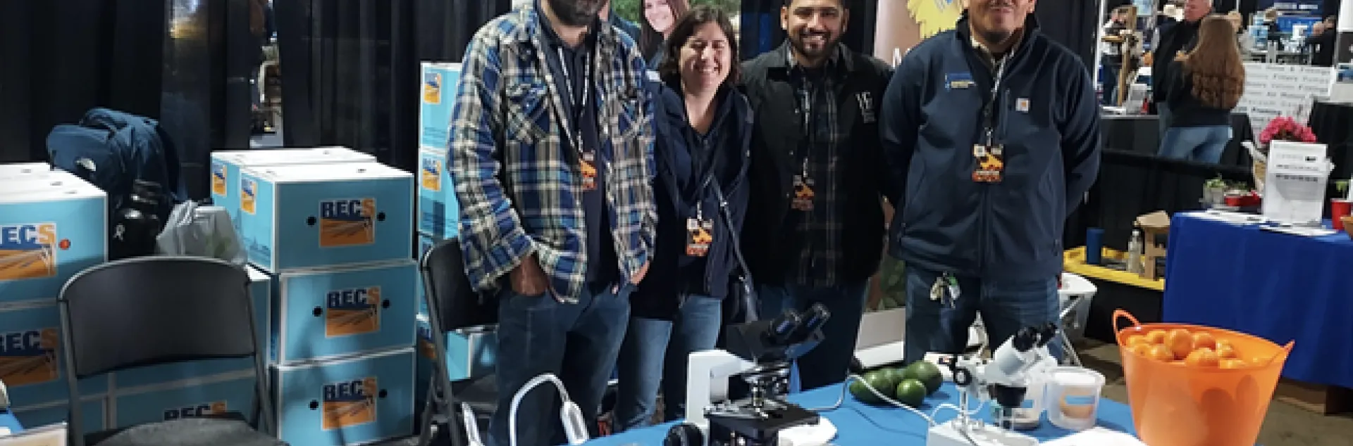 Four people stand behind a table covered with a blue tablecloth labeled Kearney Agricultural Research Extension Center and next to boxes labeled RECs