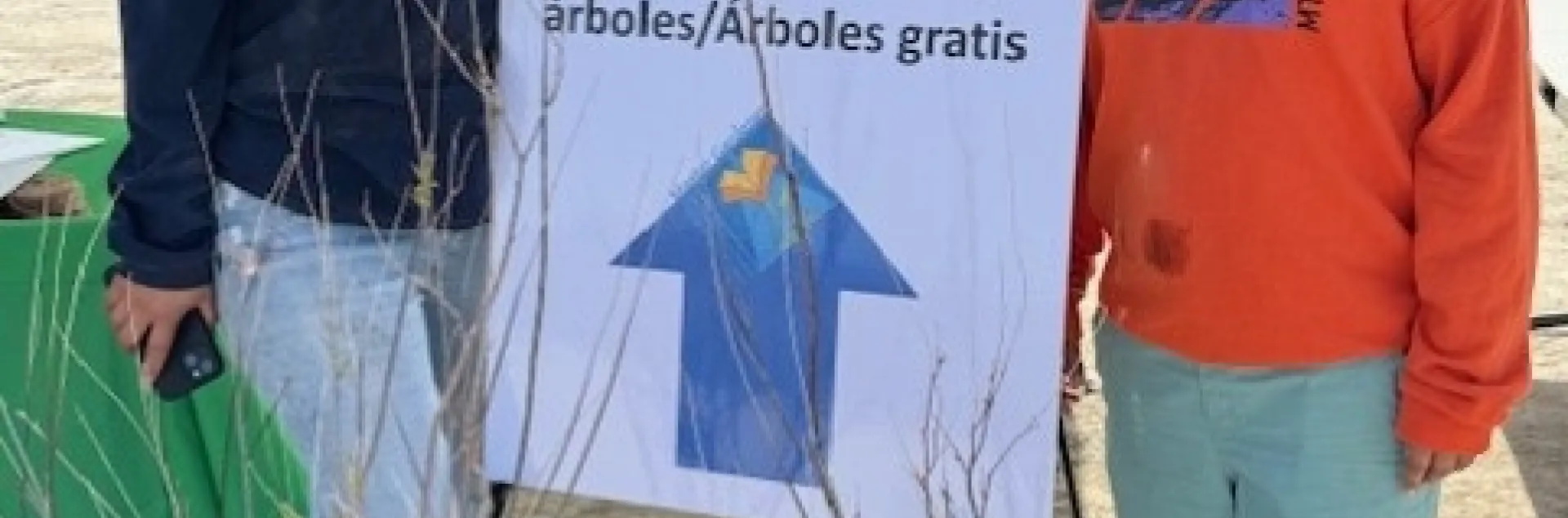 A woman and boy stand beside a sapling and sign that reads: Tree education/free trees. Educacion sobre los arboles/arboles gratis.