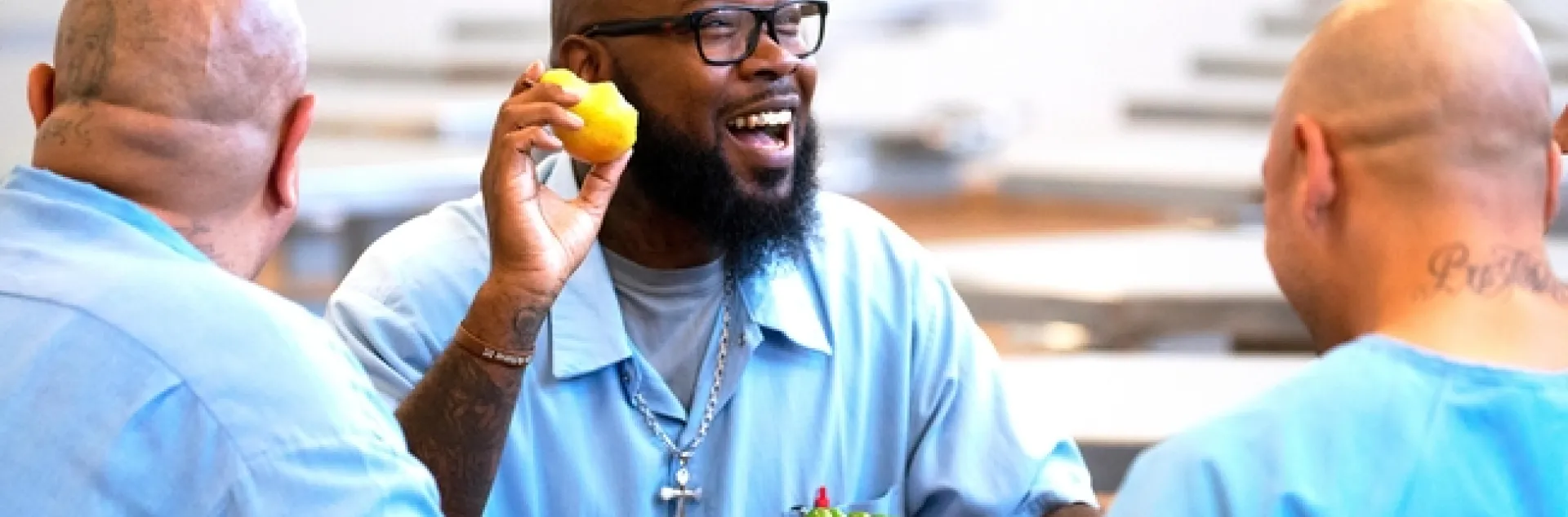 Three bald men wearing blue prison shirts sit at a table eating lunch.