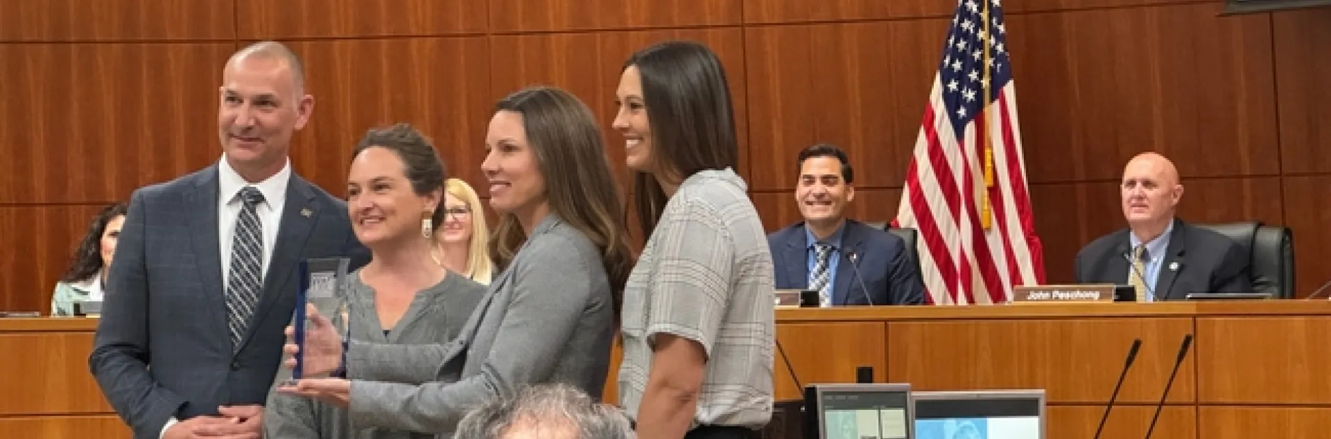 Four people pose for a photo with Shannon holding up the lucite trophy as the board of supervisors look on in the background.