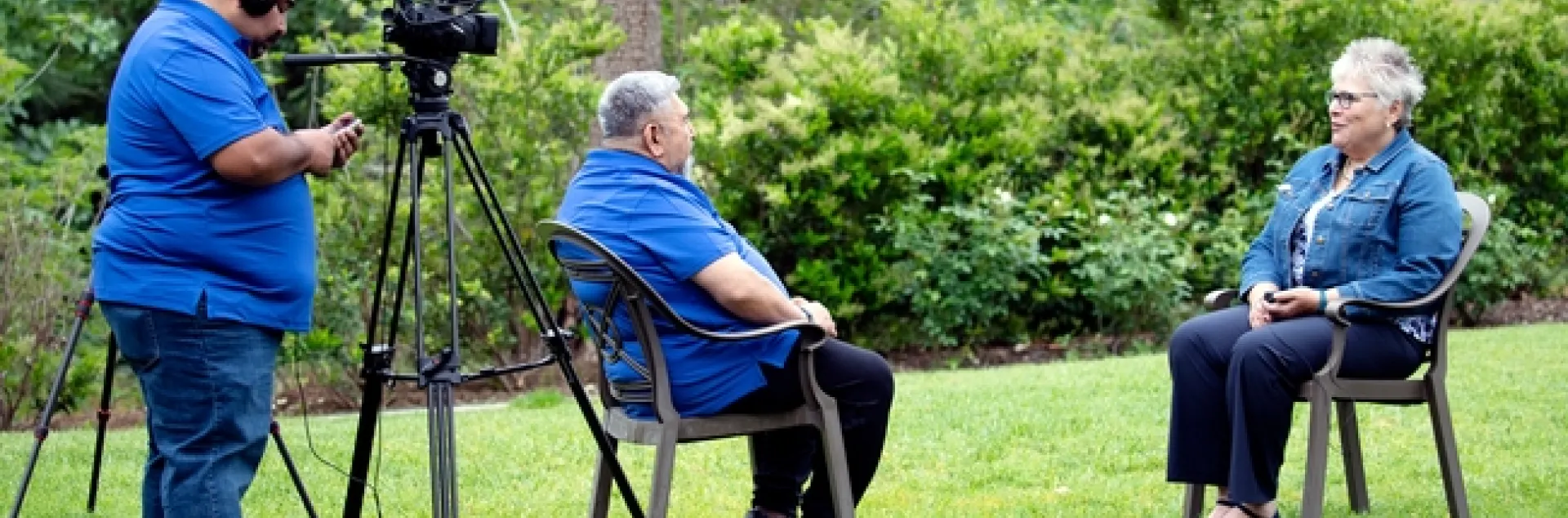 A man standing behind a camera records another man interviewing a woman while sitting down.