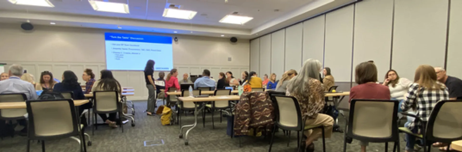 Dozens of people sit at conference room tables. PowerPoint screen reads: "Turn the Table" Discussion. Ask your BP Team questions. Jepardy topics: Procurement, T&E, C&G, Fiscal Close. Choose 2-3 topics, discuss a pain point, effciency and kudo.