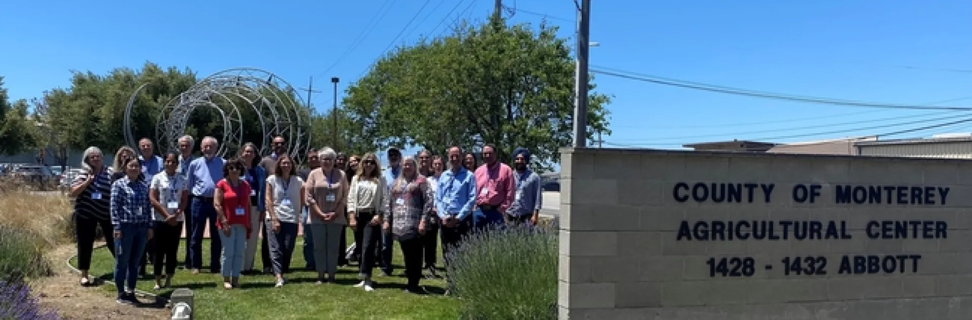 A group of a couple dozen people pose outdoors next to the "County of Monterey Agricultural Center 1428-1432 Abbott" sign.