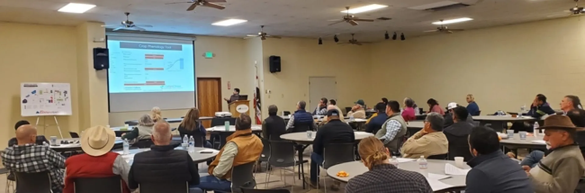 A room full of mostly men sitting around round tables look at a PowerPoint slide as Tapan Pathak describes a crop phenology tool.