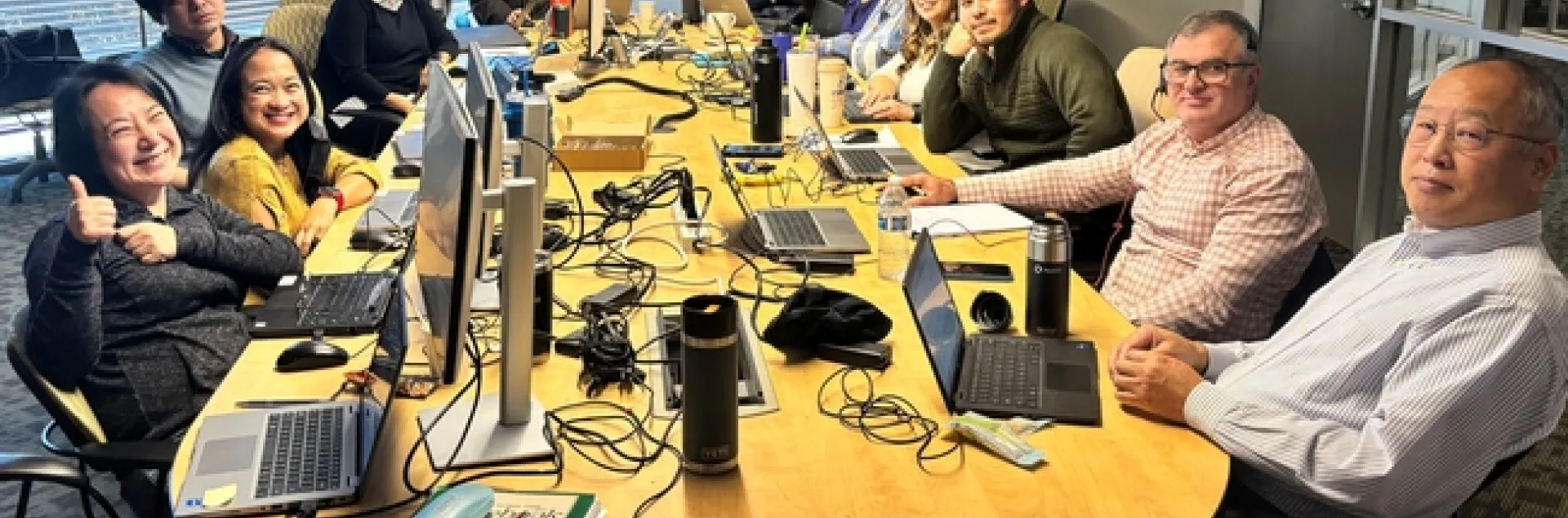 In a conference room, 14 people sit around a conference table littered with laptops.