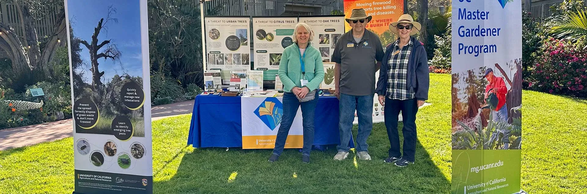 Three Master Gardeners standing in front of an informational table on green grass 