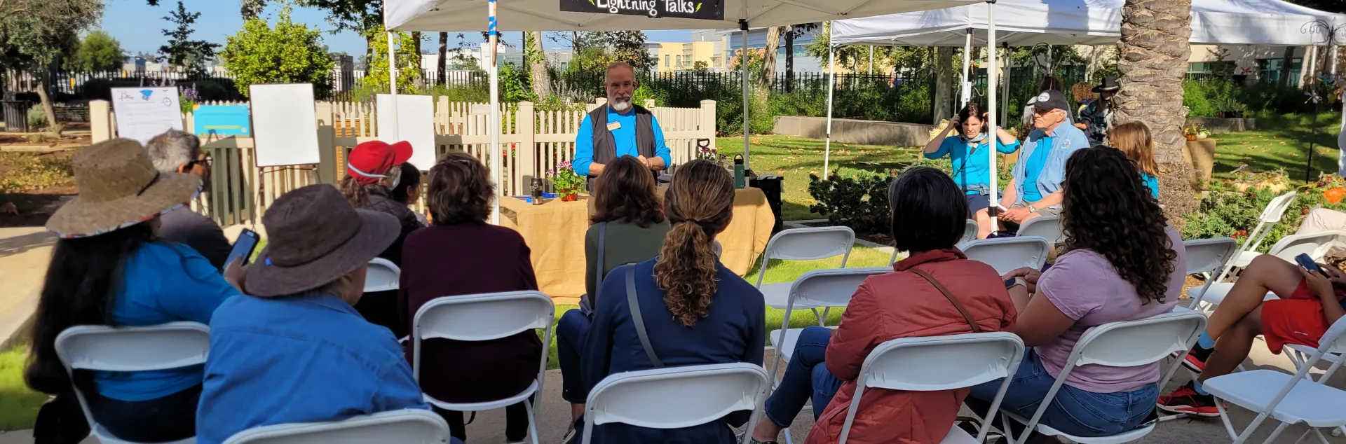 A Master Gardener under a shade with a sign that says "Lightning Talks" is speaking to an audience outdoors with onlookers nearby and a garden in the background.