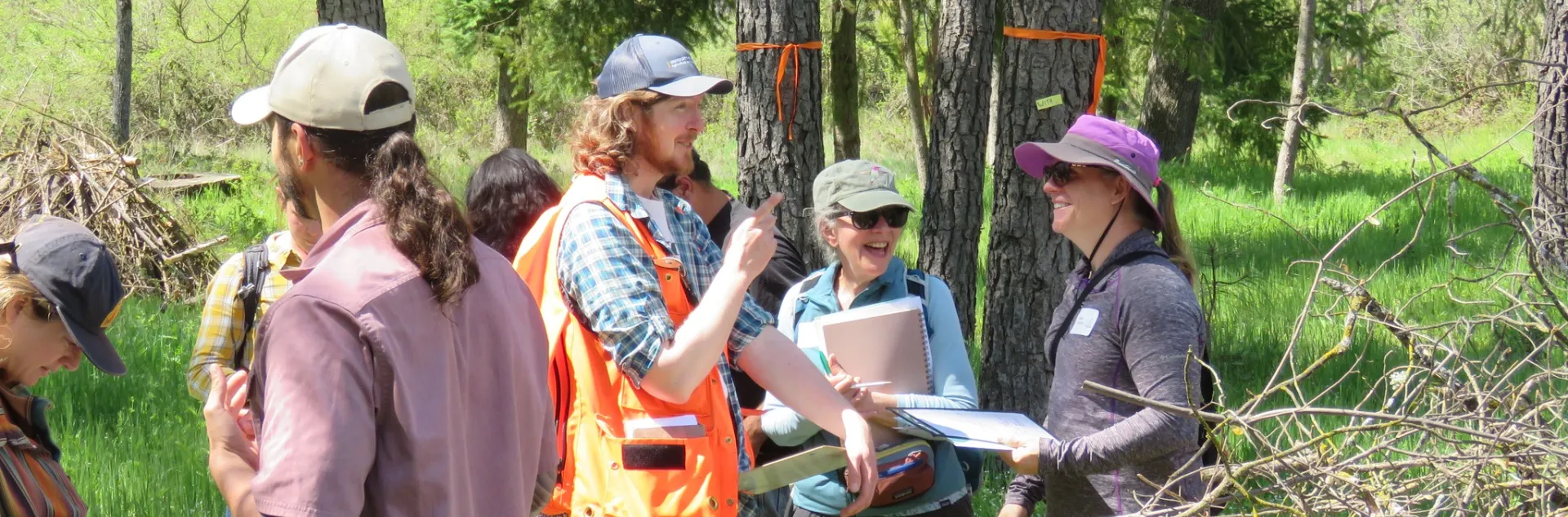 Man in cap and orange vest points to a cluster of trees as two Tree School participants laugh and smile while holding class papers and materials