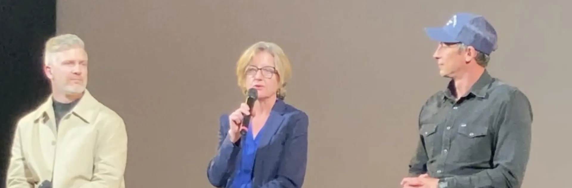 A woman seated on a stool onstage speaks into a mic as men on either side of her watch her speak
