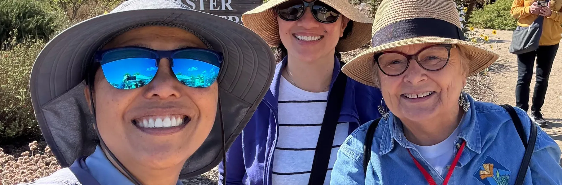 Three smiling people in wide-brimmed sun hats pose for a selfie in front of a UCCE Master Gardener wooden sign outdoors.