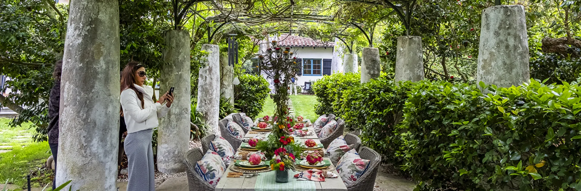 A long outdoor dining table is set with bright plates and linens underneath a lush, vine-covered metal pergola. The structure has thick weathered stone pillar legs. A woman in a white jacket and grey wide-leg trousers stands near a pillar, holding her phone to take a photo. Grey wicker armchairs with floral patterned cushions flank the table. The path is paved with large rectangular stones and box hedges, vining plants, and trees surround the area.
