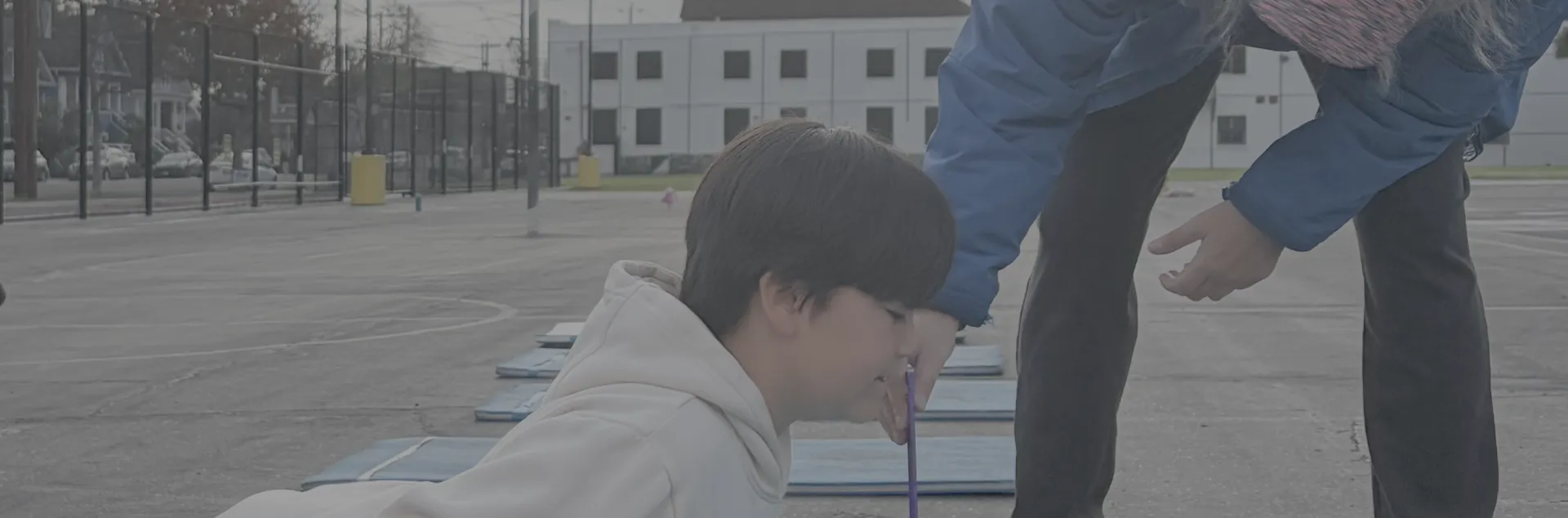 An elementary school student lays on his stomach on a yoga mat outside on the school blacktop, stretching his head and chest up while his PE teacher holds a ruler under his chin.