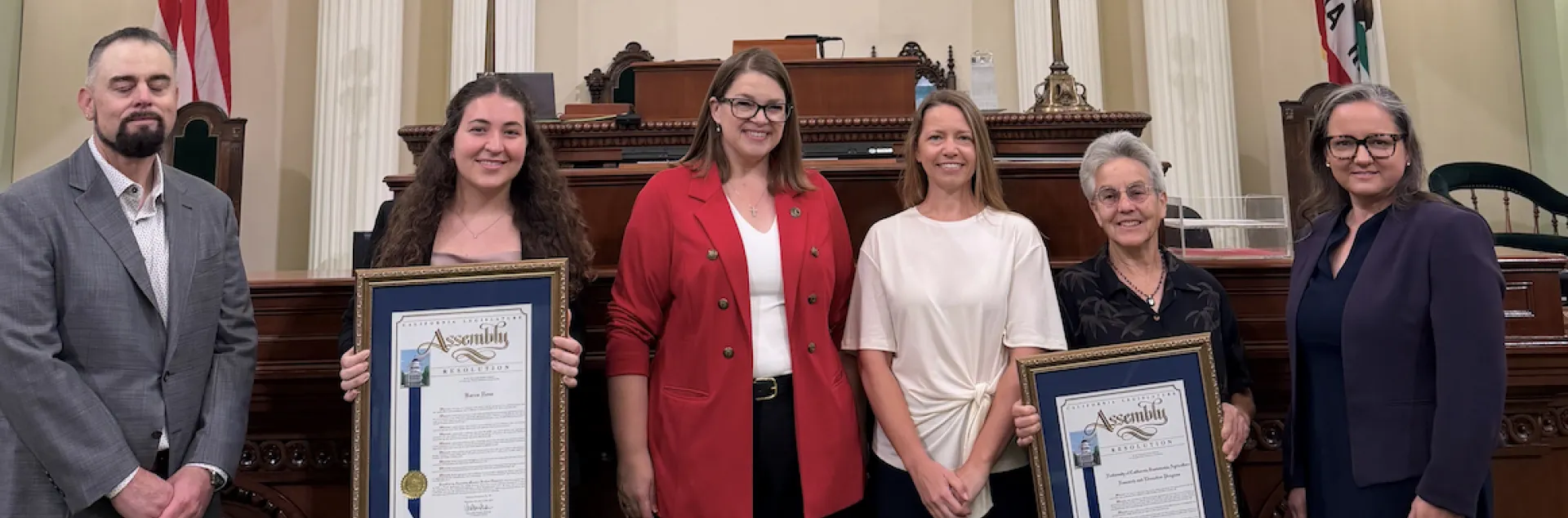 Six people stand in front of U.S. flag in the Assembly chamber. Two of the women are holding framed resolutions.