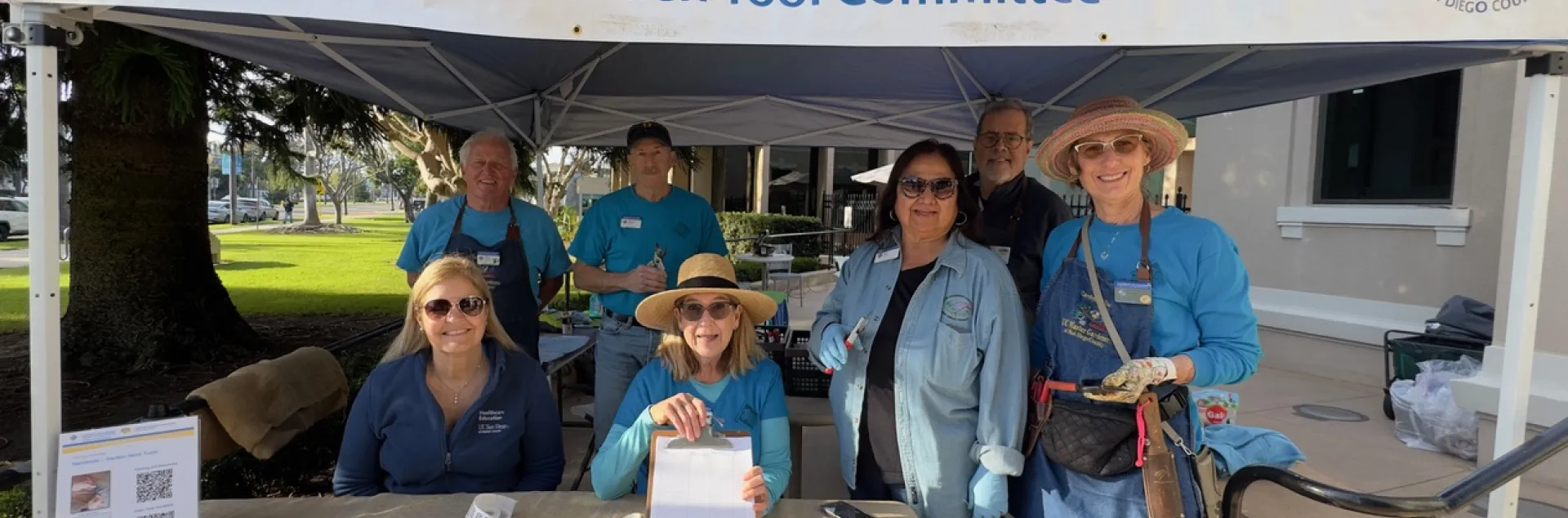 Seven people smile from under a blue canopy and white sign that reads "University of California Agriculture and Natural Resources, UCCE Master Gardener Program of San Diego County, Garden Tool Committee." The table in front of them is covered by brown burlap and has papers and a clipboard. One person is holding garden tools.