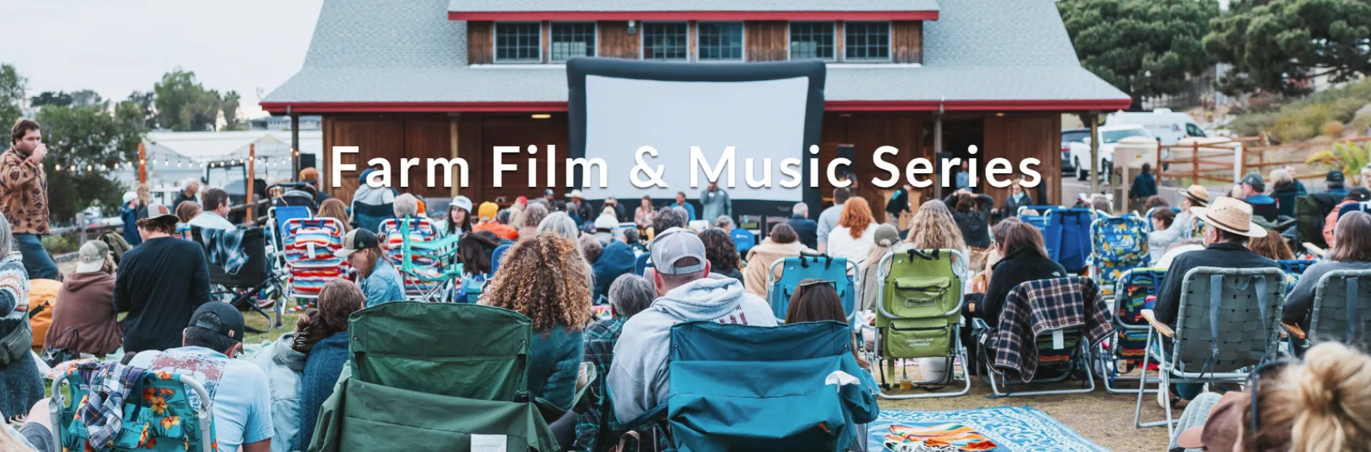 A large crowd sits in chairs facing a large outdoor movie screen set up in front of a barn-like building.