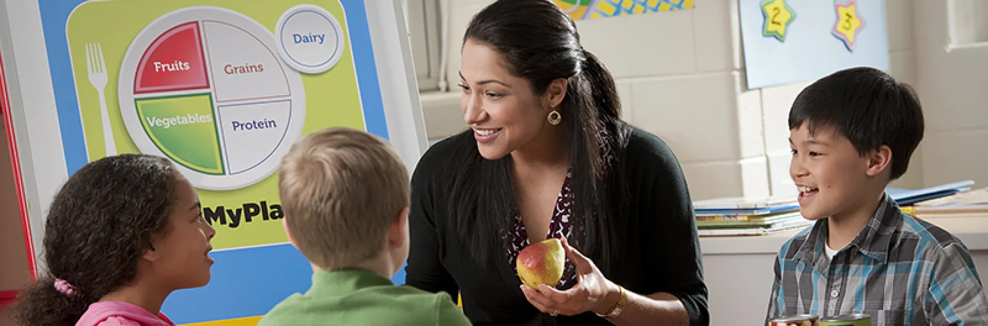 A teacher sits at a table with elementary-school aged students and talks with them about healthy eating, while holding a piece of fruit in her hands.
