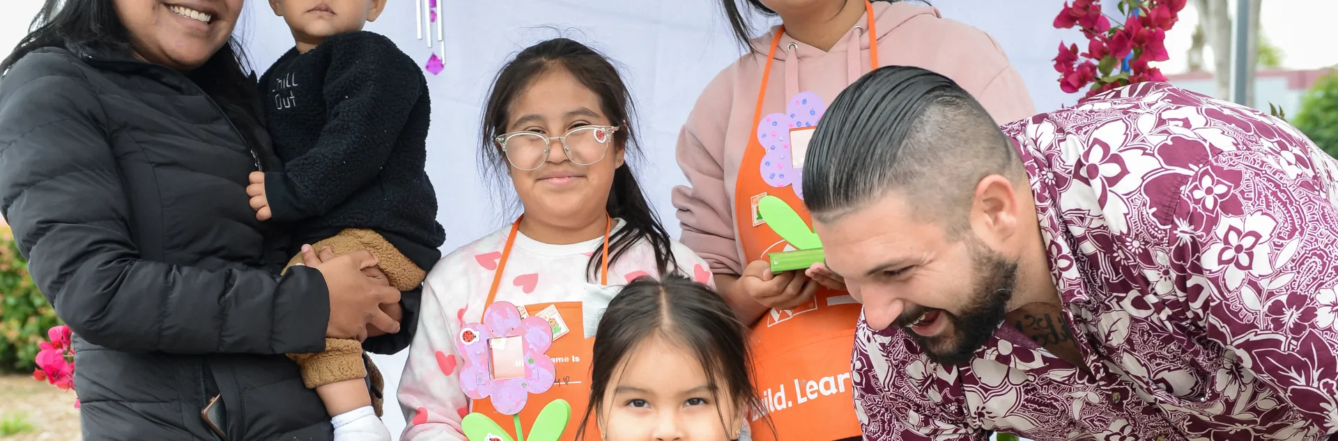 A family smiles while standing in a booth with artistic colors in the background. The family of six shows three of the four children wearing Home Depot orange aprons and holding floral crafts.