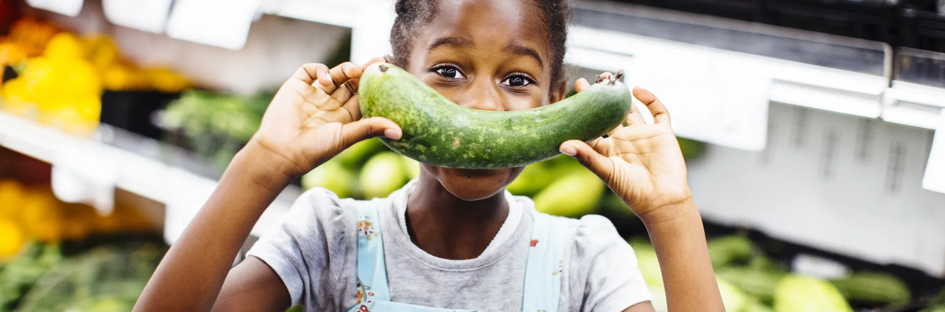 A young girl holds a cucumber in front of her face while standing in a grocery store produce aisle.