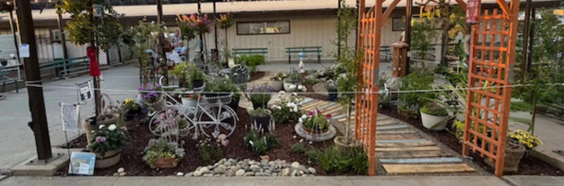 2026 Merced County Fair Demo Garden with white bicycle planter on left, teak arbor on the right, pond on right rear corner, two large white square planters in middle, iron cafe table and 2 chairs in middle left, and 50 plants laid out around these elements