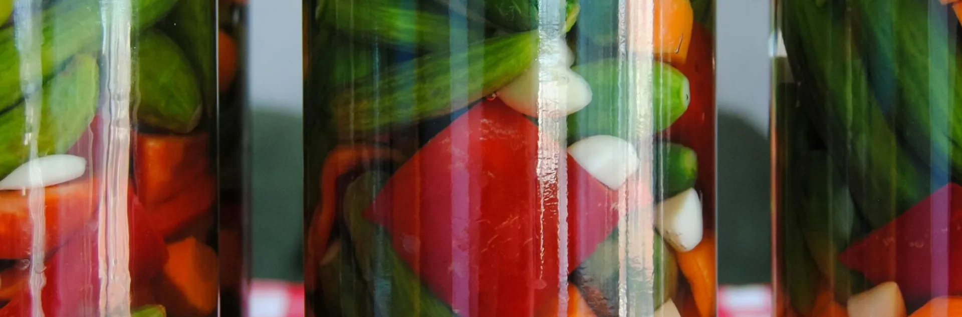 Three glass jars filled with pickled cucumbers, carrots, peppers, and garlic sit on a red and white checkered tablecloth