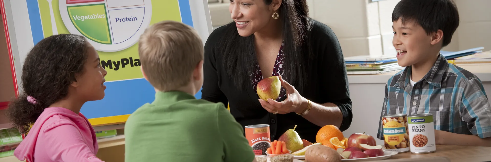 A teacher sits at a table with elementary-school aged students and talks with them about healthy eating, while holding a piece of fruit in her hands. The MyPlate graphic is visible on a poster in the background.
