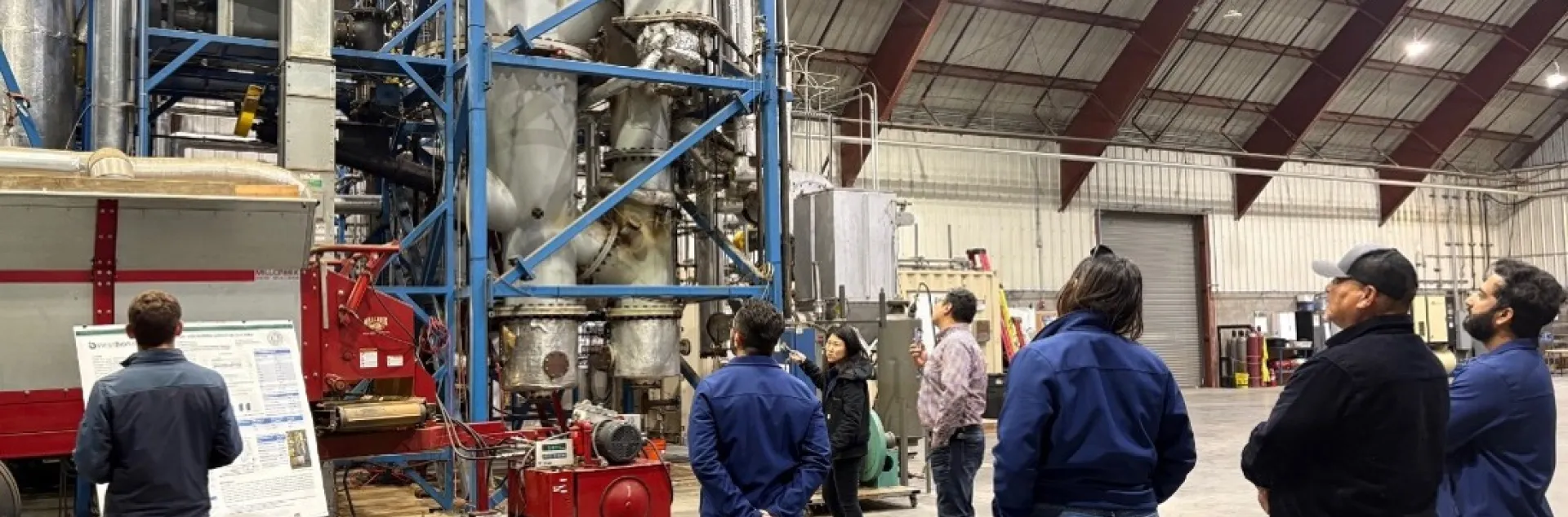 a group of people in a large industrial space look at a system of large pipes in a scaffolding about 50 feet high while someone explains the system