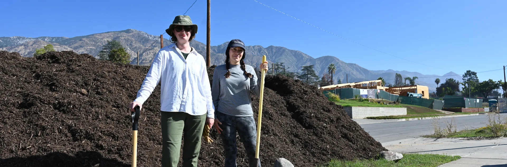 Two people with shovels stand in front of a pile of compost.
