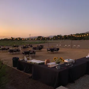 A sunset view of the outdoor annex area set up with round table and chairs and buffet, with string lights and flowers, for an event