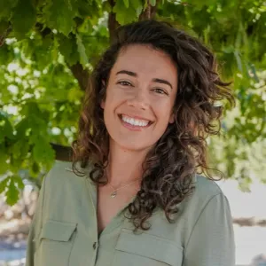 Kelsie Matteucci is wearing a green collared shirt and has curly brown hair. She is surrounded by green trees. 