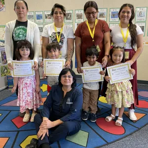 4 parents standing behind 4 children who are proudly displaying their EFNEP graduation certificates. EFNEP Educator sits on the carpet in front of them.