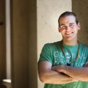 a man wearing a green shirt with his arms crossed leaning against a pillar
