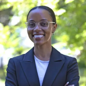 Crystal is standing in front of trees and has on glasses, a dark blazer, and a white shirt.