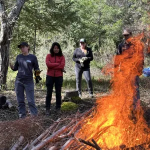 Fire Advisor, Tori Norville, leading a pile burn workshop. She is standing next to a pile of vegetation that is on fire. 