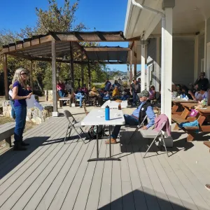 A speaker presenting to a group of community members that are sitting in front of her. They are all outside on the deck of a building.
