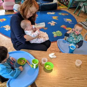 A childcare provider holds an infant in her arms while bottle feeding her. Two toddlers sit next to her in high-chairs eating snacks.