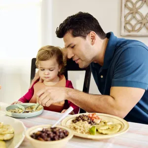 A father helps his toddler eat dinner.