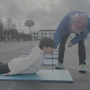 An elementary school student lays on his stomach on a yoga mat outside on the school blacktop, stretching his head and chest up while his PE teacher holds a ruler under his chin.