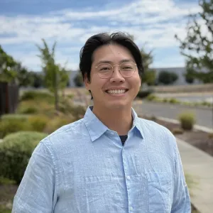 A person wearing round glasses and a light blue button-up shirt smiles outdoors against a blue sky and landscaped background.