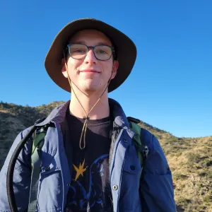 a man wearing a blue jacket, glasses, and a bucket hat standing outside in front of a hill
