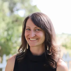 Photo of the face and shoulders of a middle aged woman with brown hair, in an outdoor setting. Hair is wavy and should length, and she wears a black tank top.