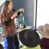 A woman with long hair, wearing a chili-patterned shirt, speaks into a microphone while holding a jar in an outdoor setting, with an audience in wide-brimmed hats watching her.